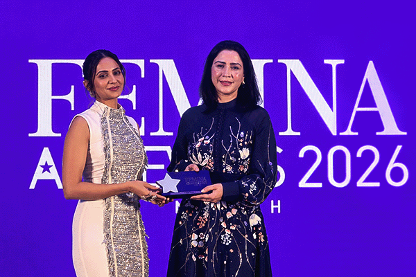 Two women on stage at the FEMINA Awards 2026; a presenter in a white dress hands a trophy to the recipient in a dark floral dress, against a purple backdrop.