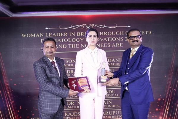 Woman in a white suit receives a trophy from two men in suits on a stage at an award ceremony.